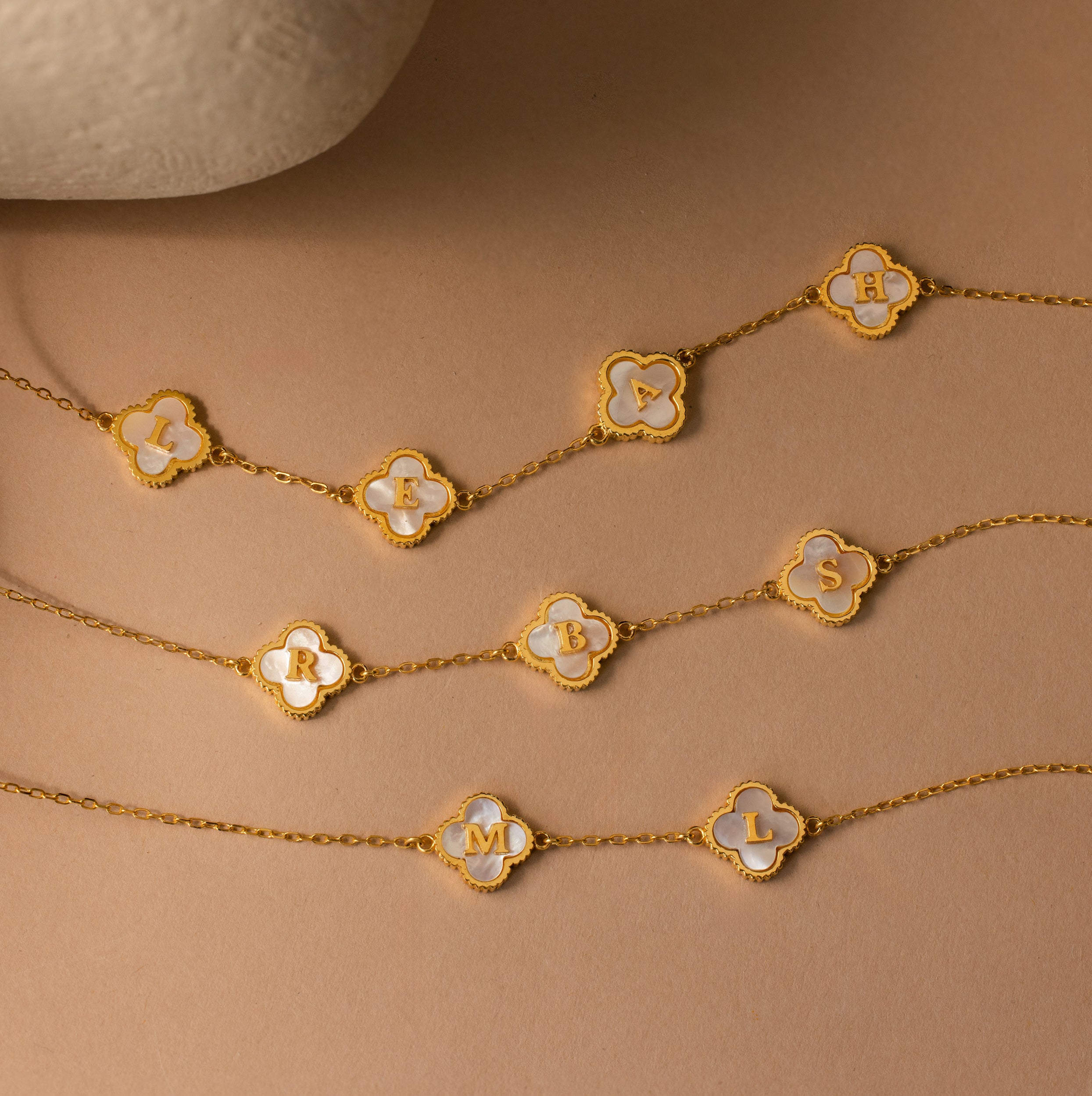 The Pearl Clover Letter Necklace features a gold chain with a clover-shaped charm and a single white letter on a beige background—a perfect personalized piece to enhance your jewelry collection.