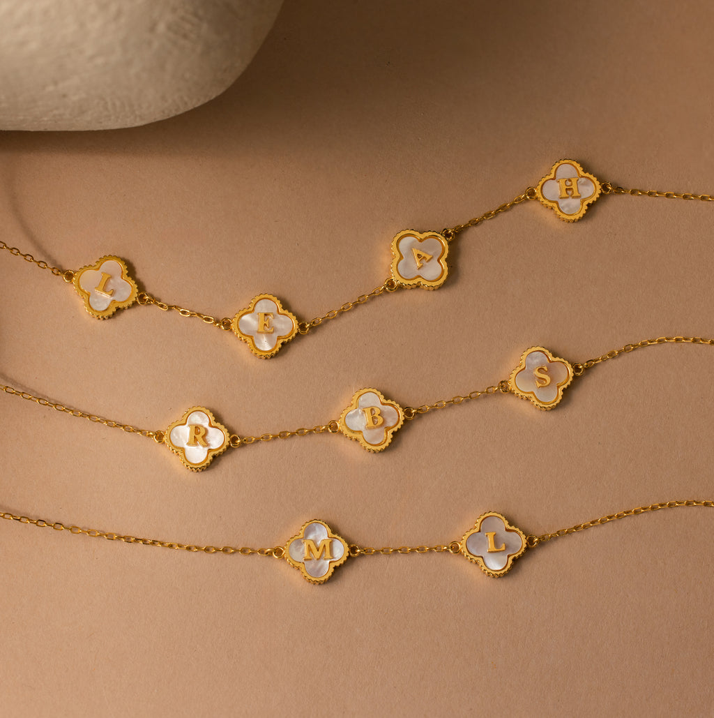 The Pearl Clover Letter Necklace features a gold chain with a clover-shaped charm and a single white letter on a beige background—a perfect personalized piece to enhance your jewelry collection.
