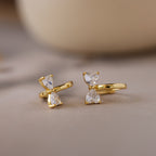 Diamond Bow Huggies featuring two vertically set pear-cut clear gemstones, displayed on a soft beige surface.