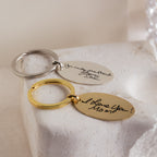 Two Handwriting Oval Keychains—one silver, one gold—featuring personalized messages are shown on a white stone surface, making each unique keychain a memorable keepsake.