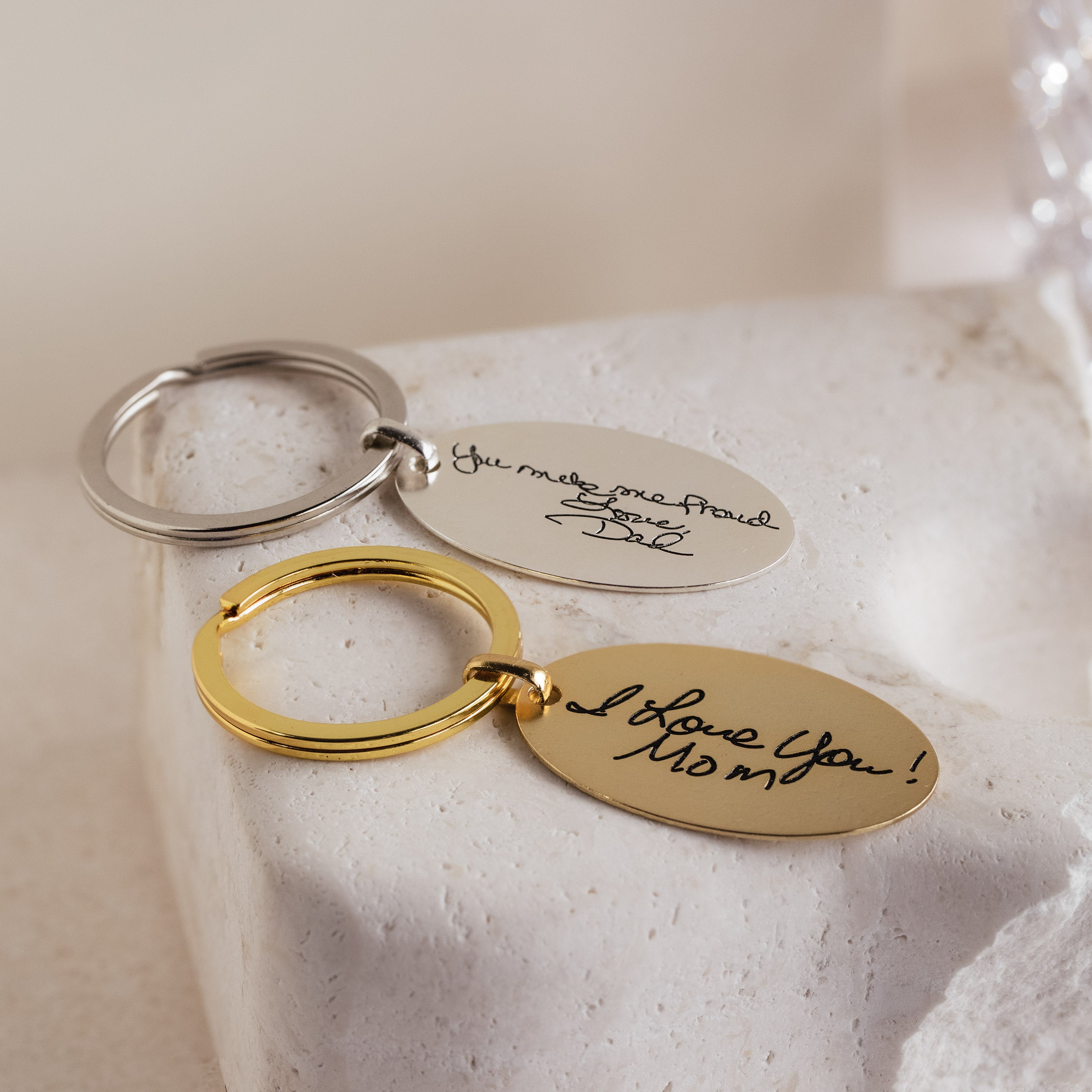 Two Handwriting Oval Keychains—one silver, one gold—featuring personalized messages are shown on a white stone surface, making each unique keychain a memorable keepsake.
