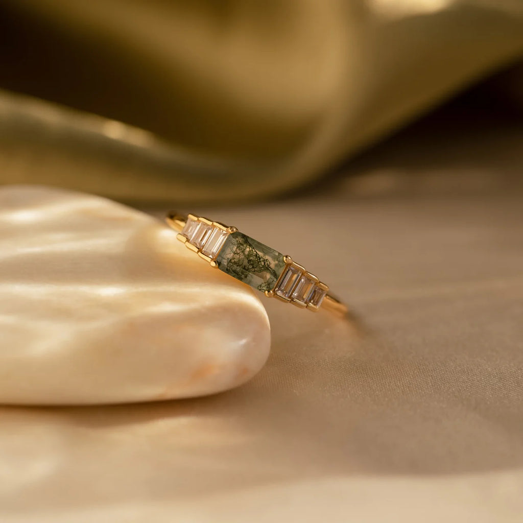 The Agate Diamond Rings Set features an Art Deco design with a green agate gemstone and rectangular clear stones, elegantly displayed on a shell against a golden fabric—an eye-catching vintage jewelry piece.