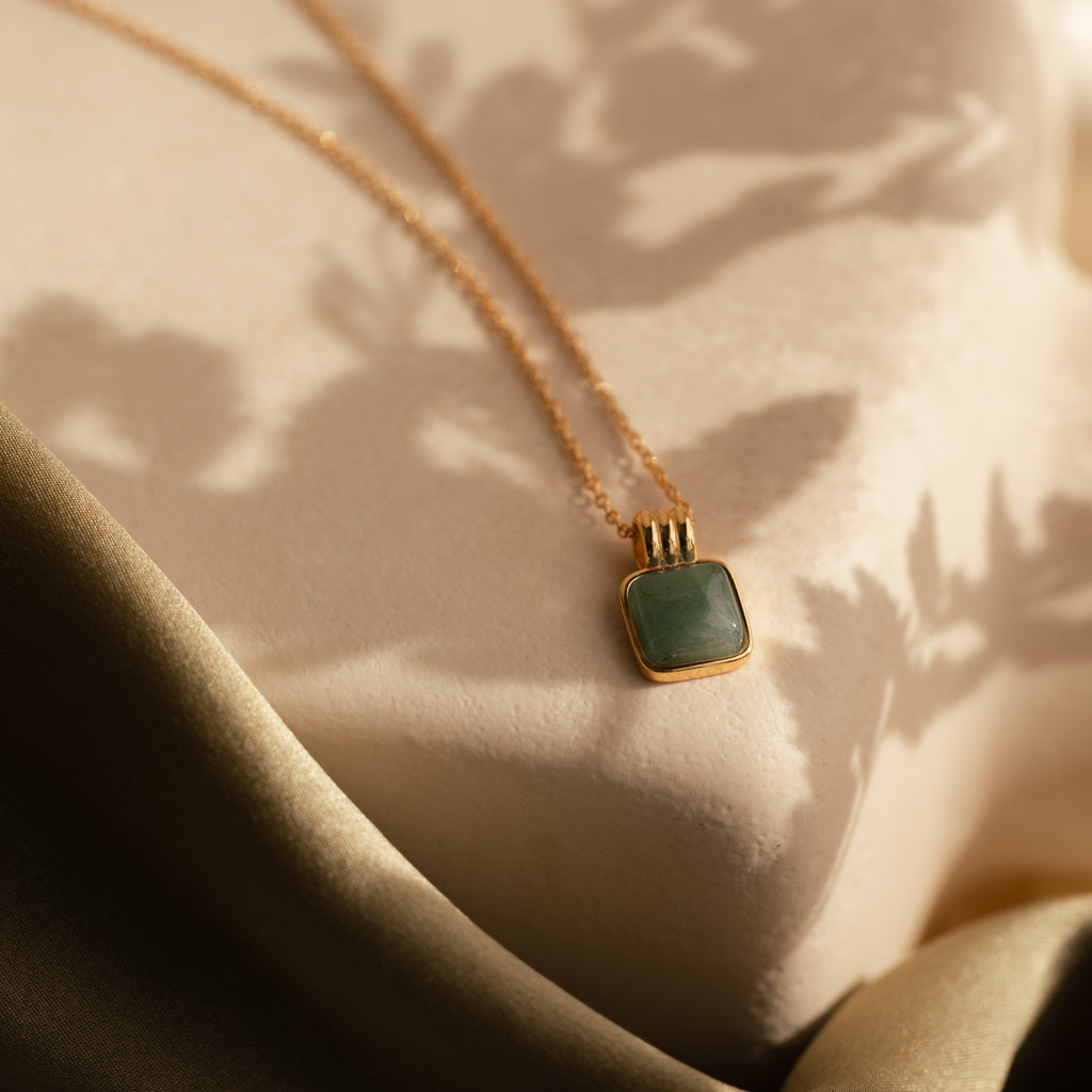 The Jade Pendant Necklace in 18K Gold, featuring a square green jade stone said to bring luck, rests on a textured surface amid soft shadows and draped fabric.