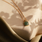 The Jade Pendant Necklace in 18K Gold, featuring a square green jade stone said to bring luck, rests on a textured surface amid soft shadows and draped fabric.