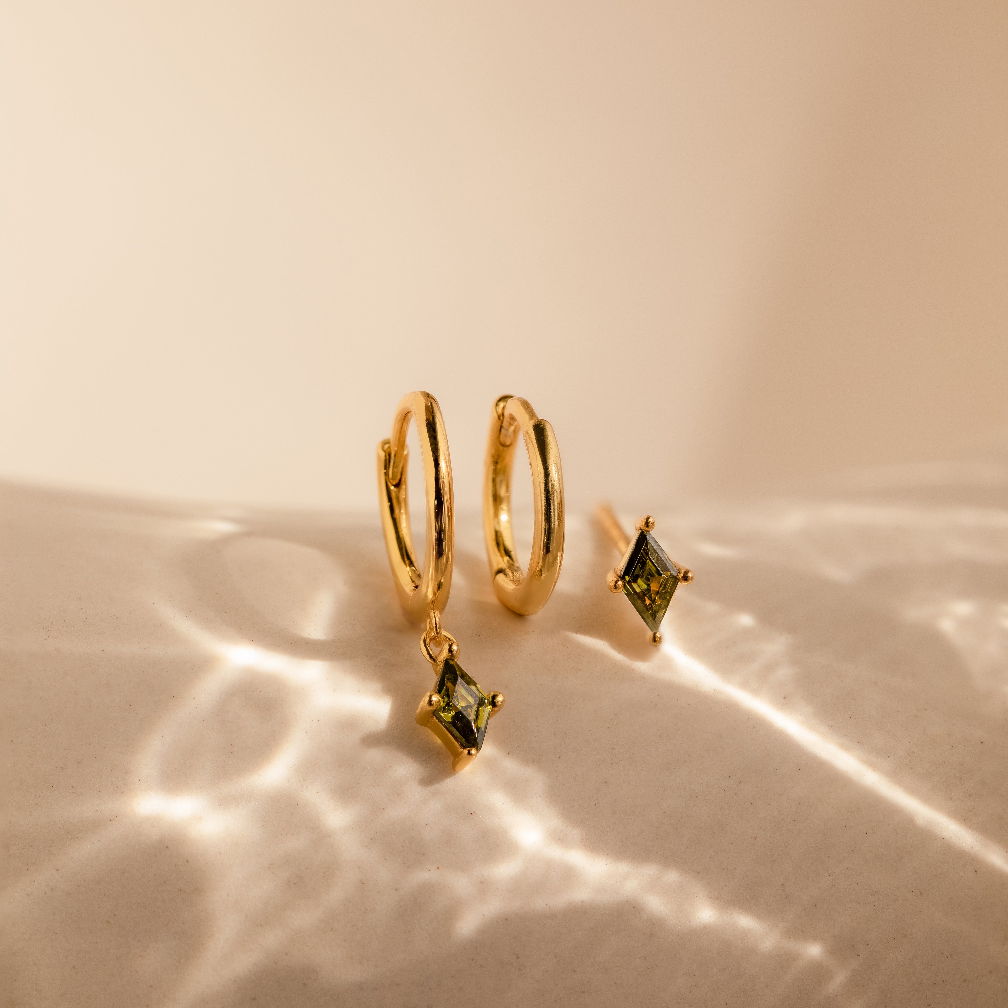 The Peridot Earrings Set features gold hoops with green peridot gemstone charms, displayed on a light, textured surface with soft shadows—perfect stacking earrings for any occasion.