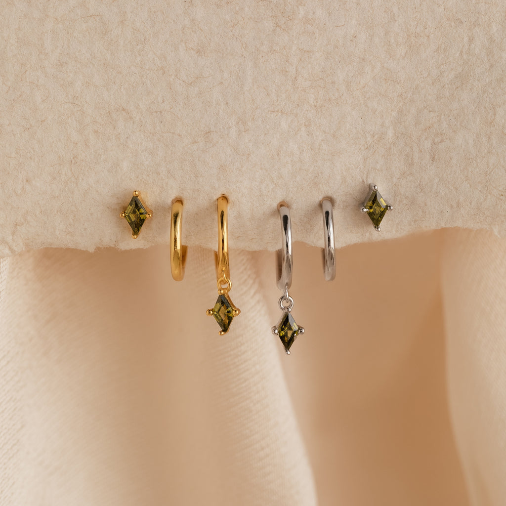 The Peridot Earrings Set in Sterling Silver features four stacking earrings with green gem charms—two gold and two silver—displayed on a soft beige fabric background.
