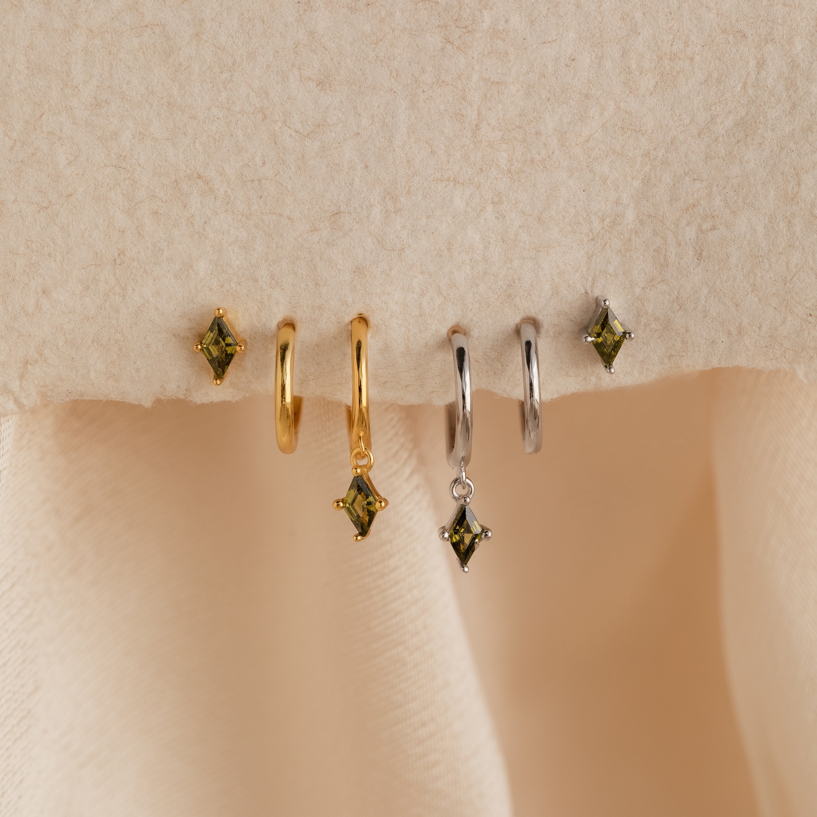 The Peridot Earrings Set in Sterling Silver features four stacking earrings with green gem charms—two gold and two silver—displayed on a soft beige fabric background.