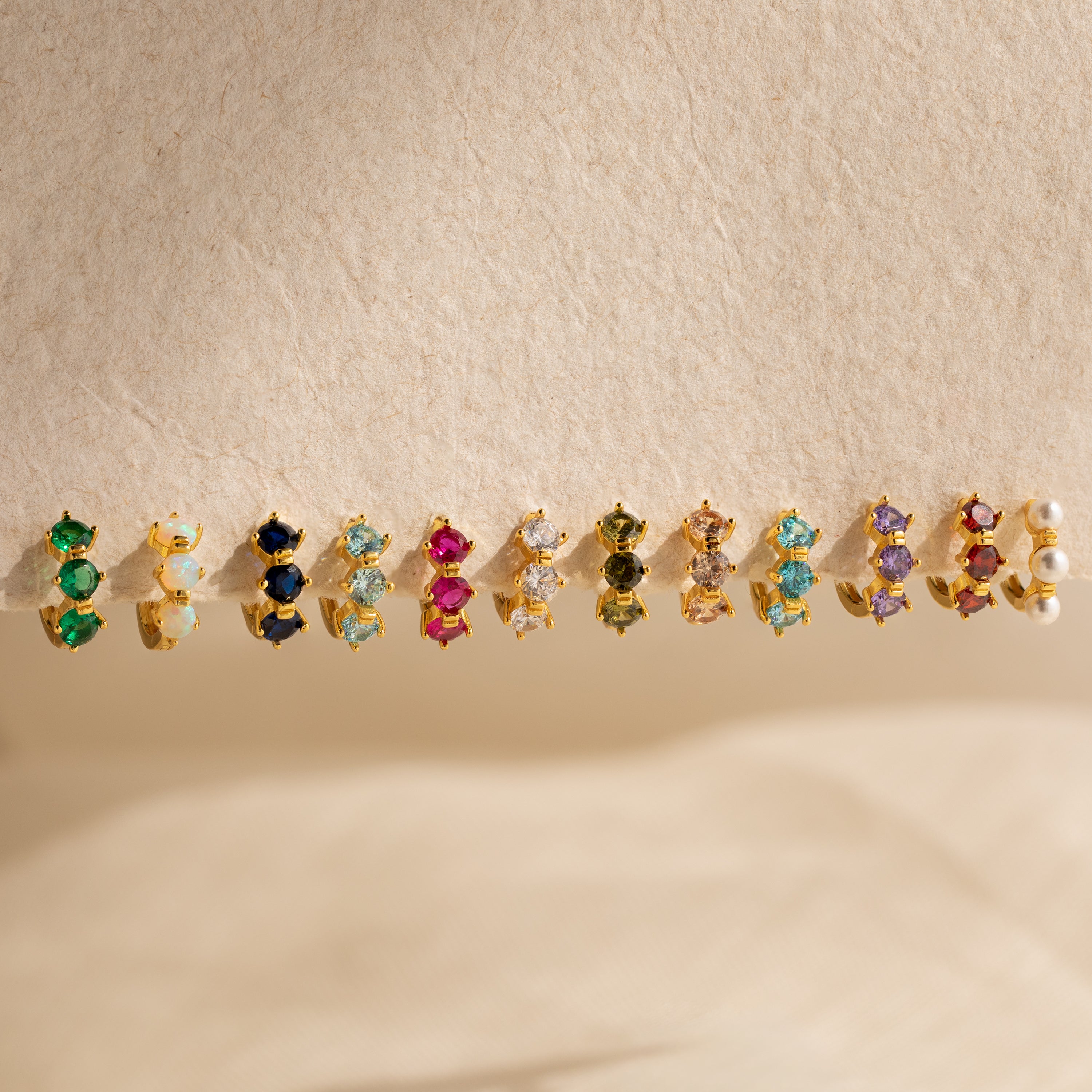 A row of Triple Peridot Huggies, featuring vibrant peridot gemstones in gold settings, displayed on a beige textured surface.