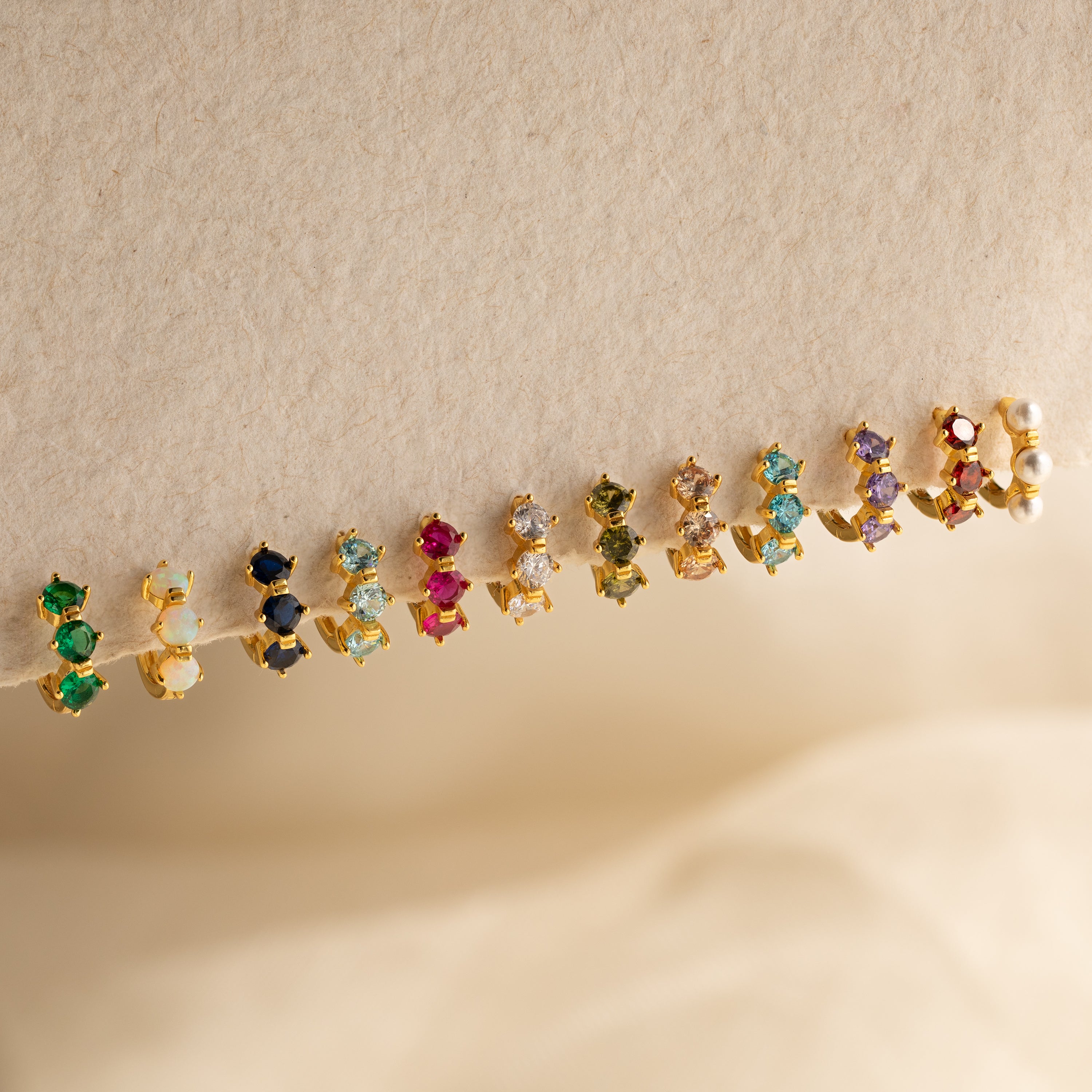 Twelve pairs of small, colorful customizable earrings, including Triple Blue Zircon Huggies, minimalist hoops, and two pearl studs are arranged on a beige background.