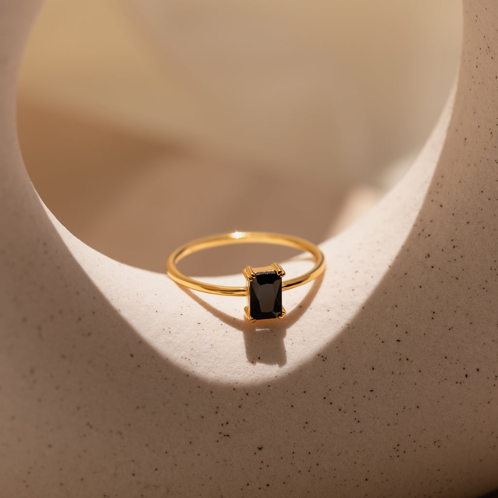 The Black Emerald Ring, a vintage-inspired gold Art Deco piece with a rectangular black stone, rests on a textured, curved white surface in soft lighting.