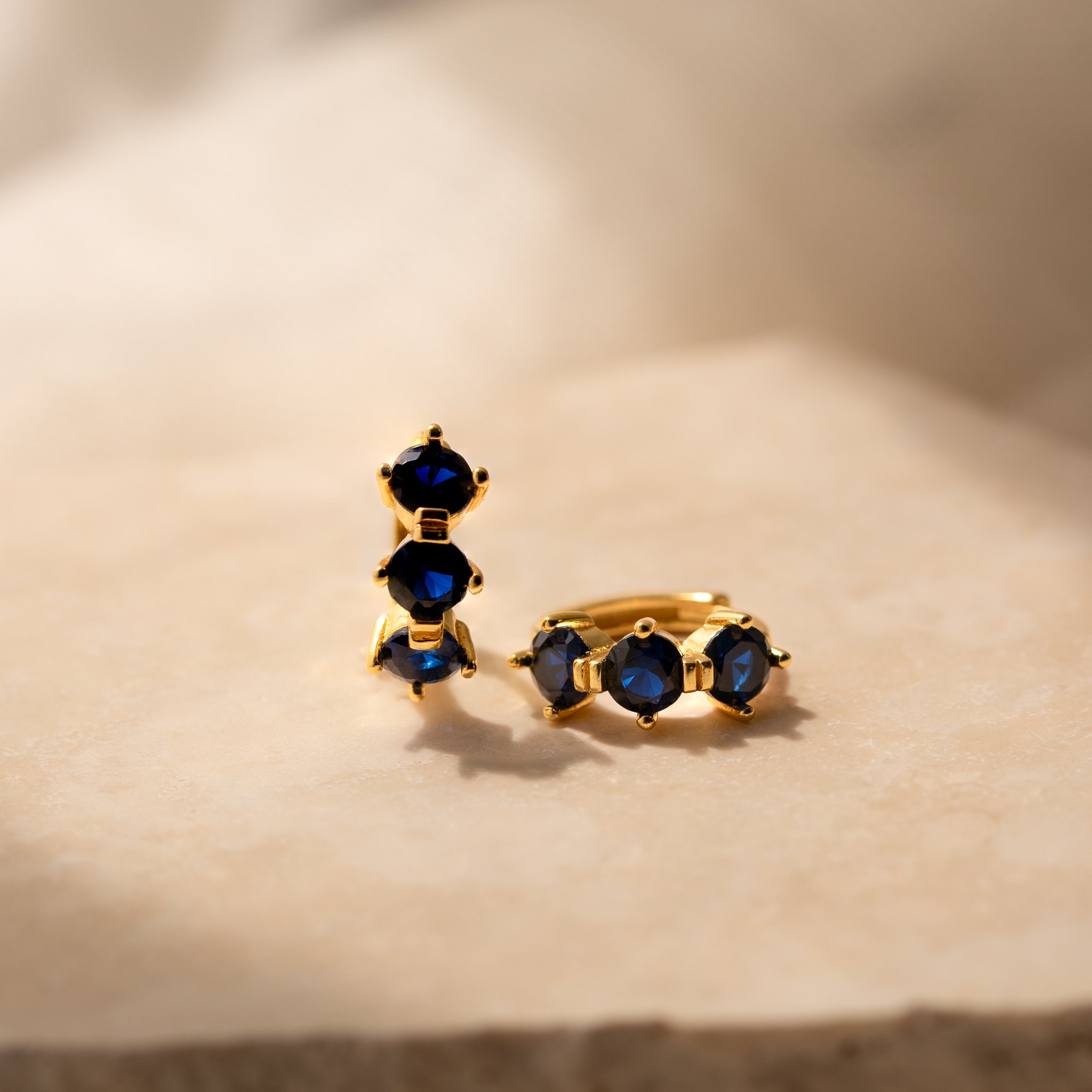 Triple Sapphire Huggies featuring gold earrings with three deep blue sapphires each, displayed on beige marble in soft light—ideal minimalist hoops for those who love personalized jewelry.