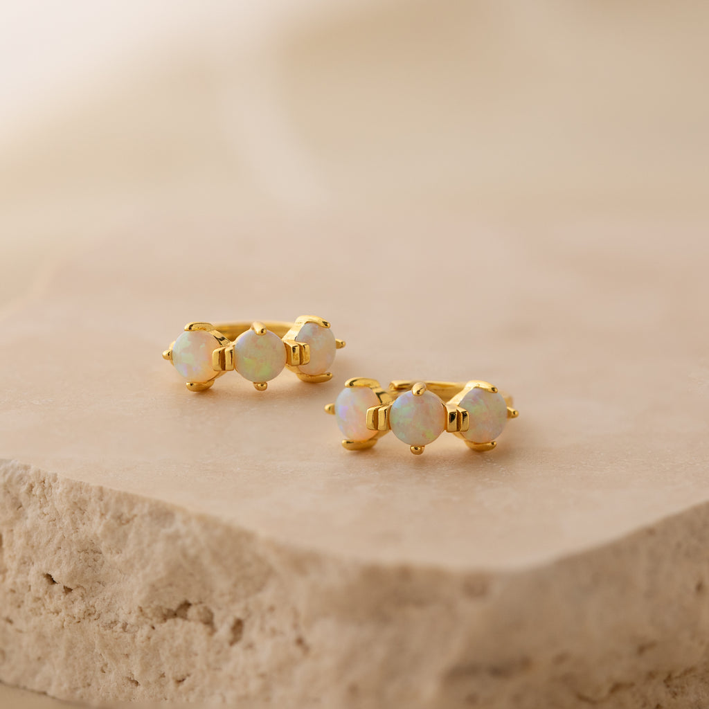 Triple Opal Huggies featuring three round opals each, shown on a beige textured stone. These minimalist hoops can be customized with your preferred birthstones for a personalized accessory.