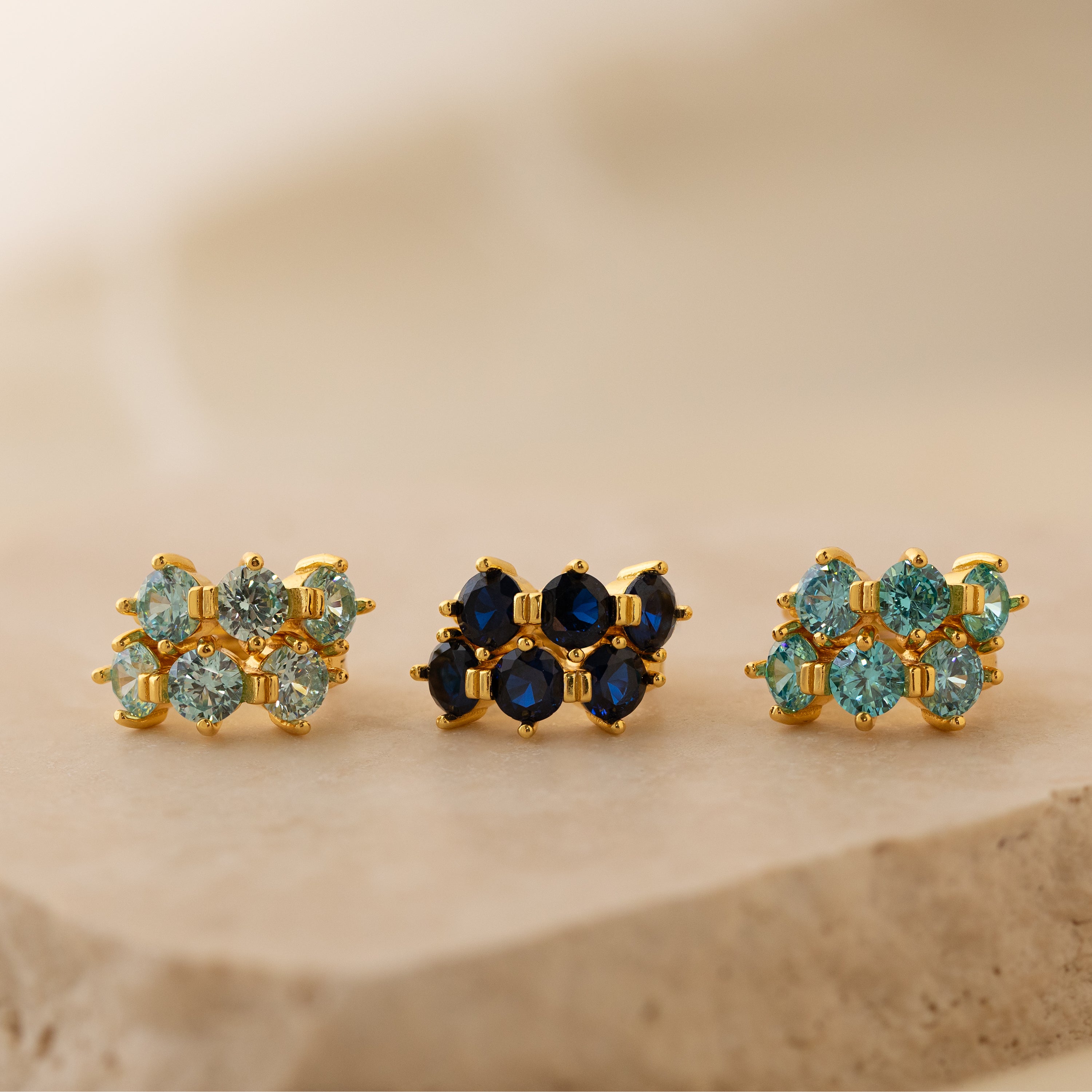 Three pairs of Triple Aquamarine Huggies earrings, featuring customizable birthstones and gemstones in blue and green hues, are displayed on a beige stone surface.