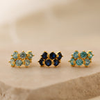Three pairs of gold stud earrings with blue and green gemstones, including the Triple Sapphire Huggies, are displayed on a beige stone surface.
