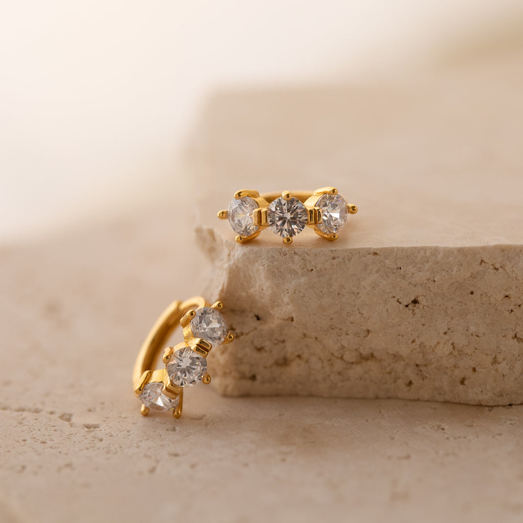 Triple Birthstone Huggies, featuring three clear gemstones on each hoop, offer customizable or unique birthstone jewelry options and are shown on a textured beige stone surface.