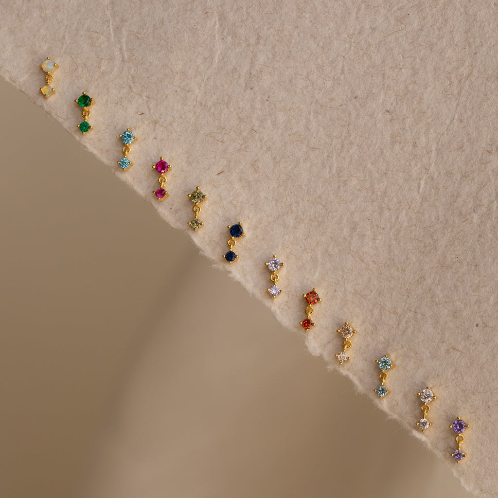 Petite, colorful Aquamarine Drop Studs are arranged in a gentle curve on a textured beige surface—ideal as birth month jewelry.