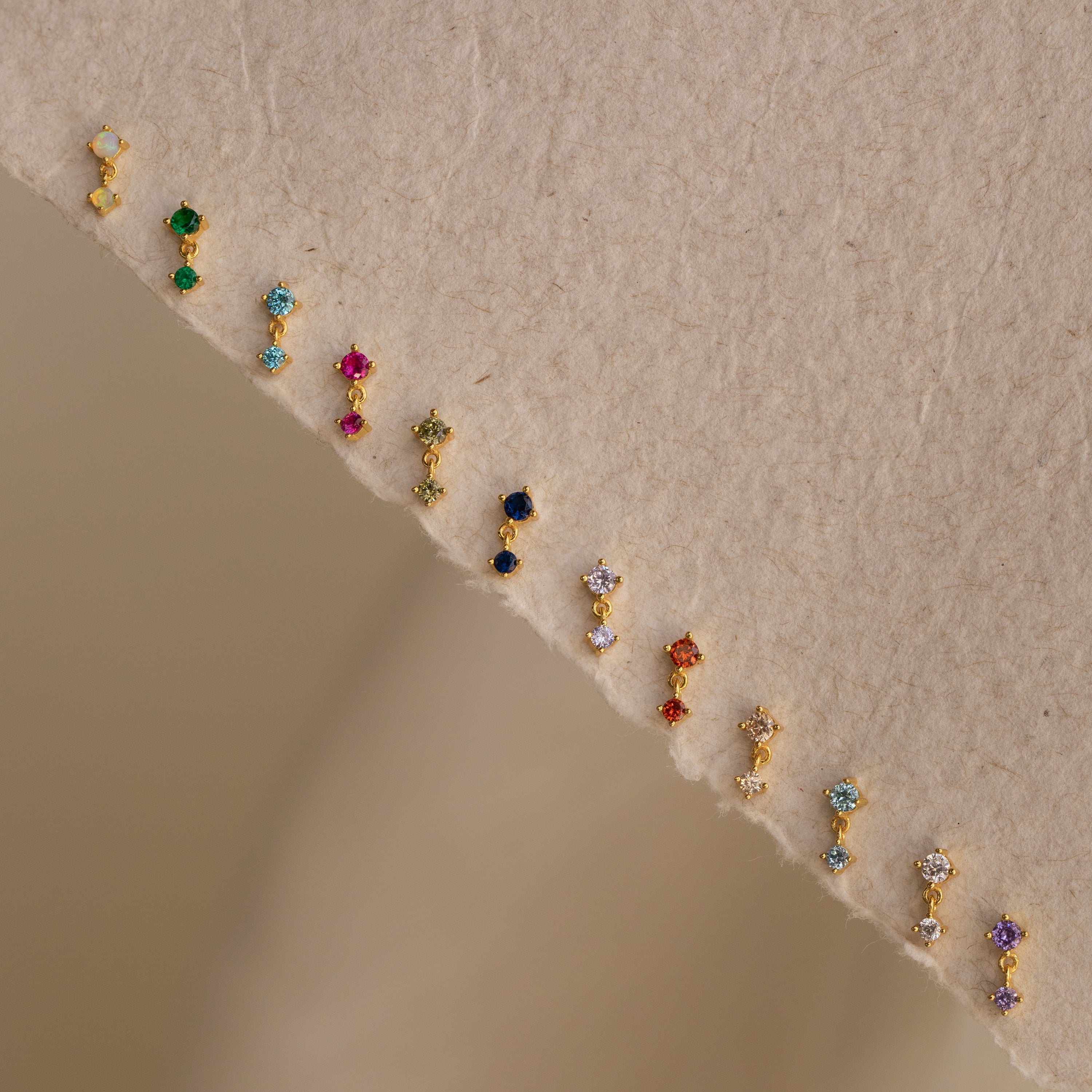 Petite, colorful Aquamarine Drop Studs are arranged in a gentle curve on a textured beige surface—ideal as birth month jewelry.