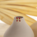 The Birthstone Drop Studs, featuring red gemstones, are displayed on a white vase with a soft beige background.