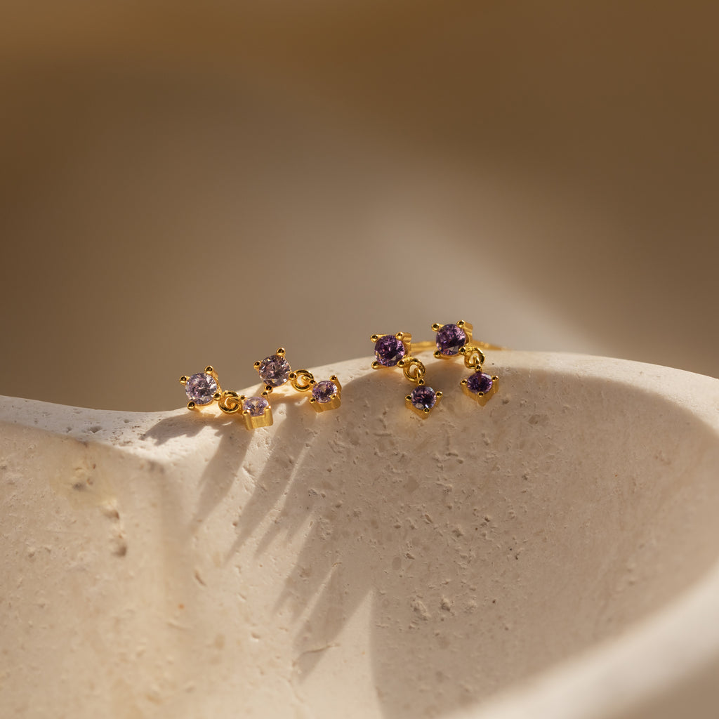 The Amethyst Drop Studs, featuring gold and purple gemstones, are birthstone earrings displayed on a textured, light-colored stone surface in gentle lighting.