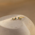Blue Zircon Drop Studs with small blue stones rest on a smooth, light-colored stone surface in soft lighting.