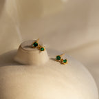 Emerald Drop Studs—gold earrings with green gemstones—are displayed on a smooth, beige surface under soft lighting.