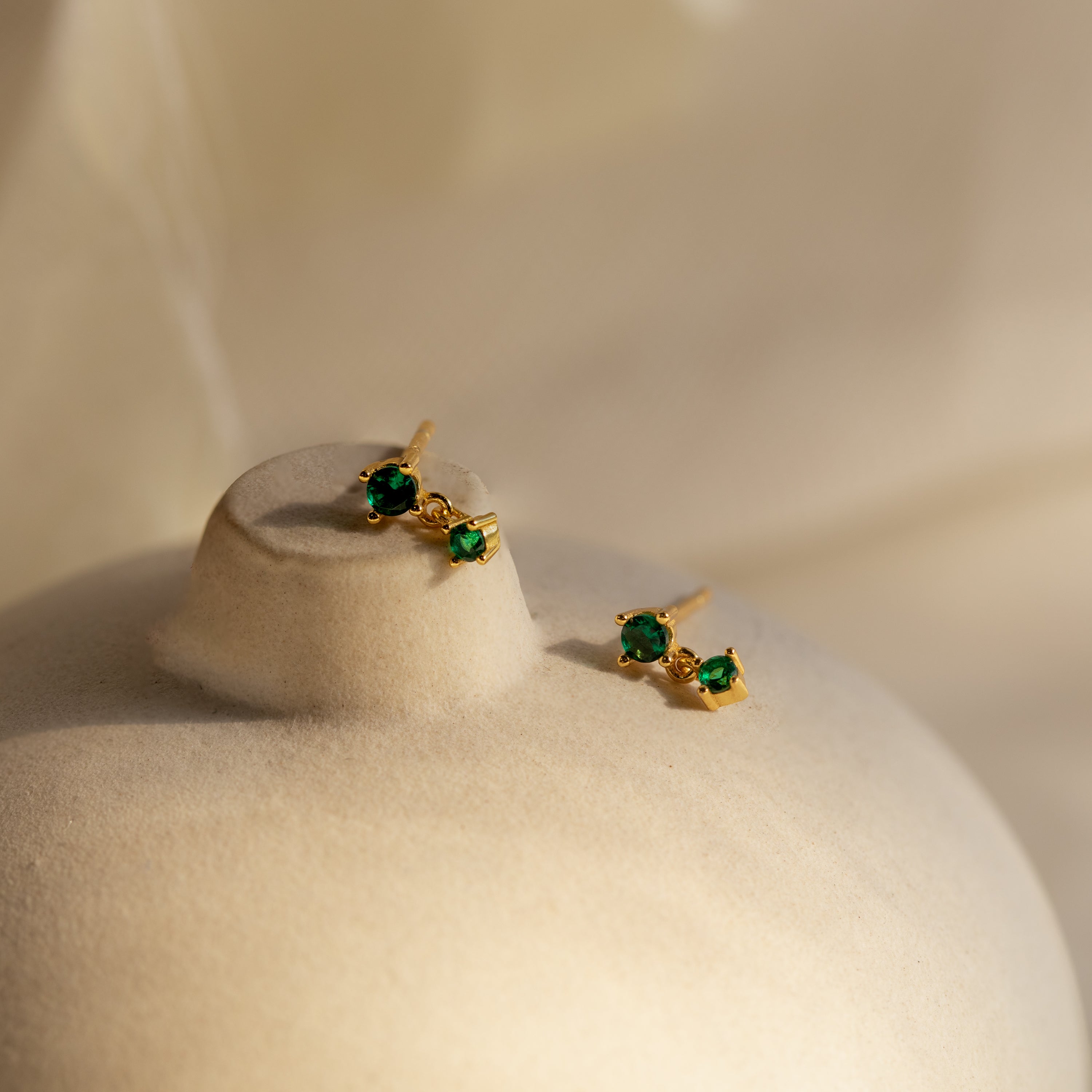 Emerald Drop Studs—gold earrings with green gemstones—are displayed on a smooth, beige surface under soft lighting.