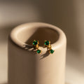 Emerald Drop Studs with green gemstones are displayed on a smooth, beige cylindrical stand under soft lighting.