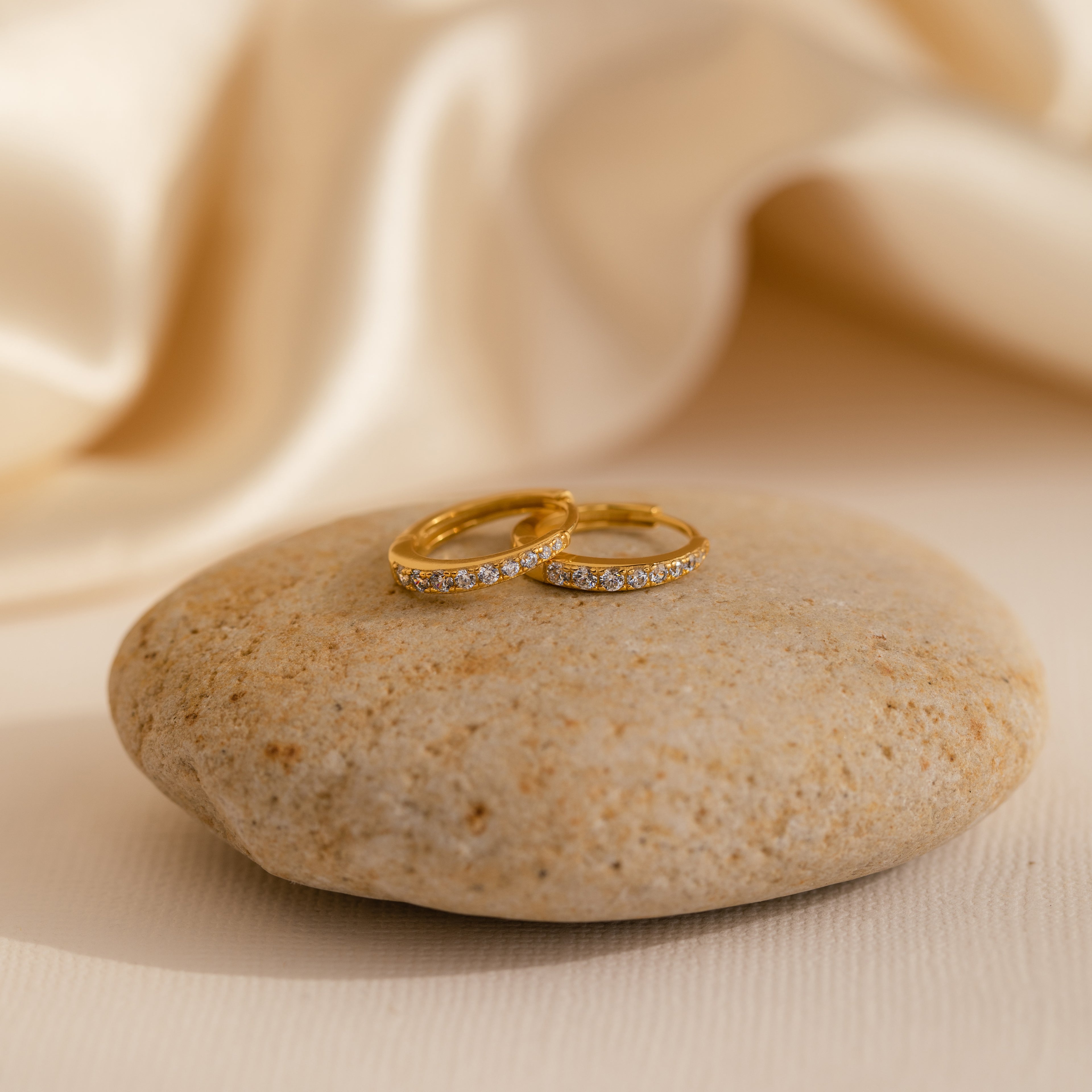 The Vera Pave Pink Tourmaline Huggies sit on a smooth stone with a soft beige fabric background, capturing elegance—perfect to pair with diamond rings for a timeless look.