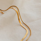 The Herringbone Duo Chain Necklace in 18K Gold rests on soft, white textured fabric with gentle, wavy folds.