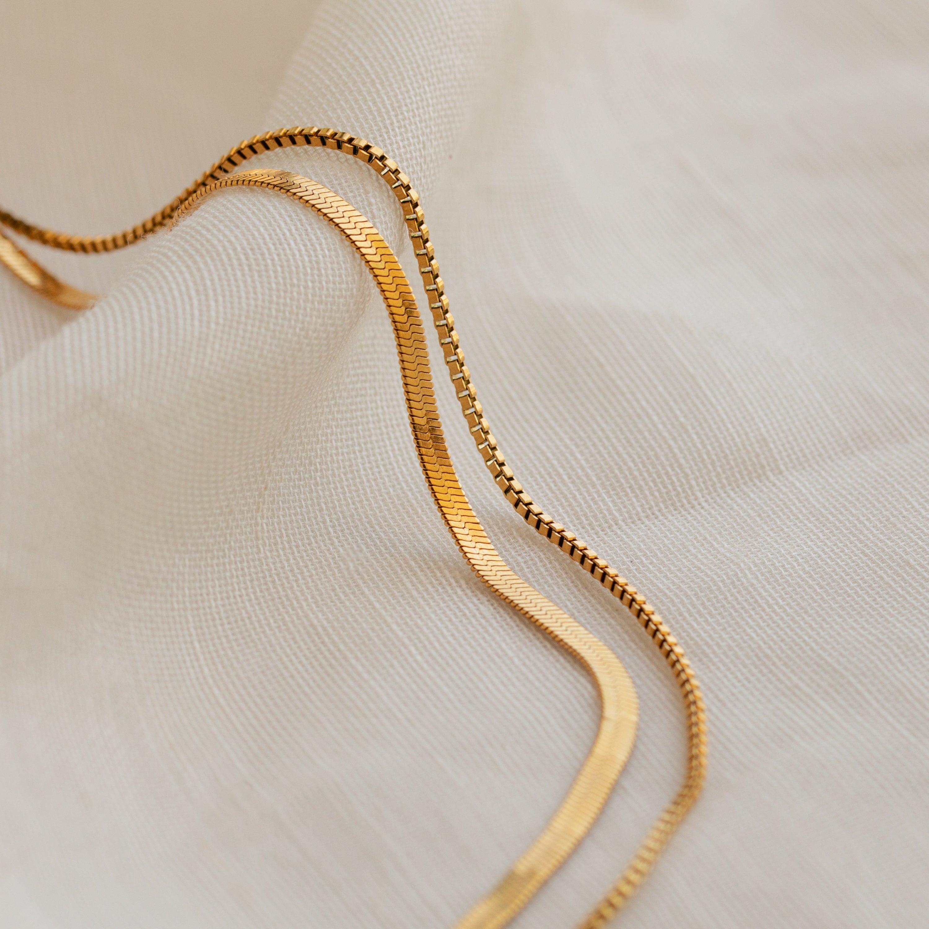 The Herringbone Duo Chain Necklace rests in soft curves on a textured fabric background, highlighting its elegant, minimalist design.