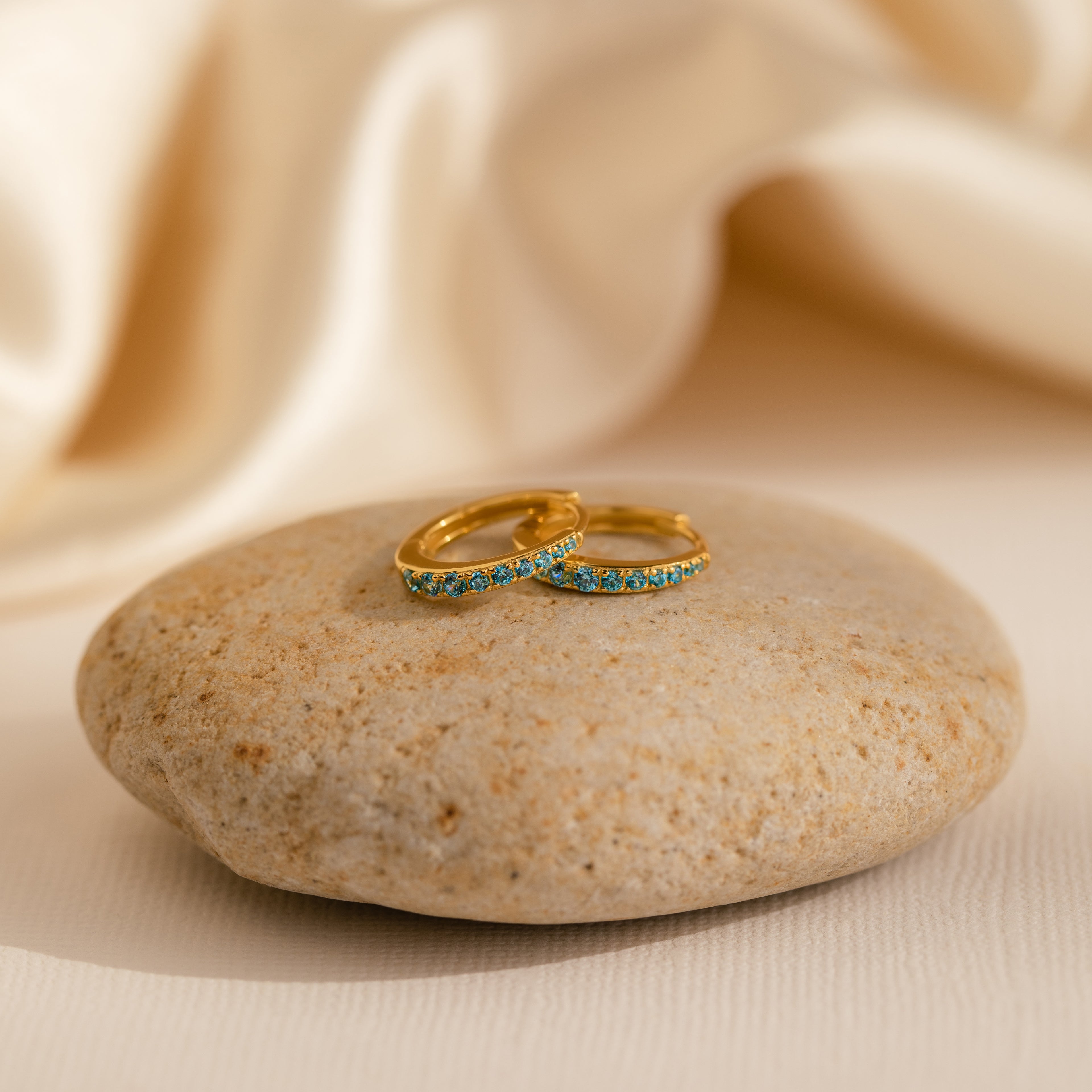 The Vera Pave Blue Zircon Huggies feature sparkling blue stones on gold hoops, displayed on a smooth stone with a soft cream fabric backdrop—ideal for adding an elegant touch or pairing with gemstone and colorful earrings.