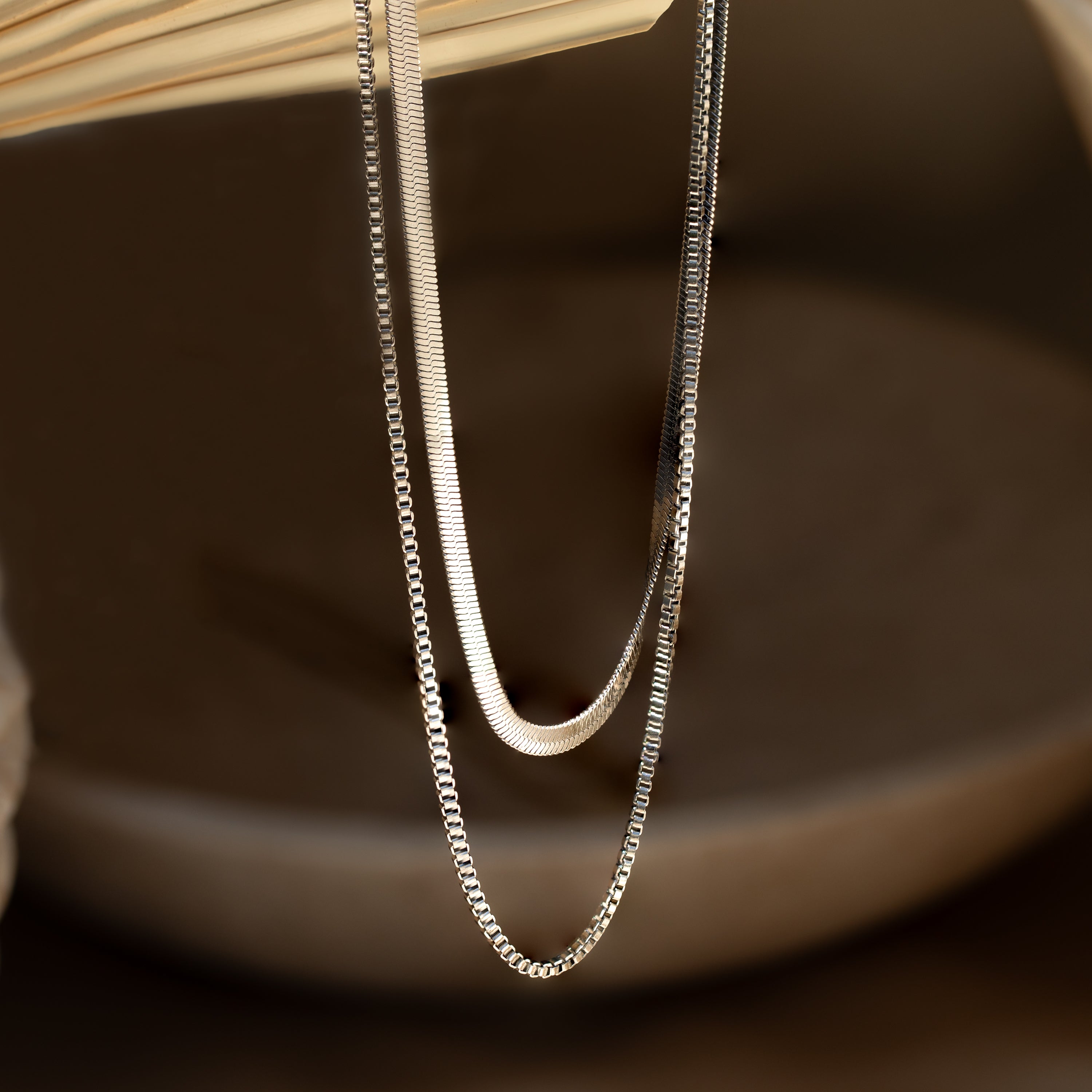 Two silver chains hang elegantly, reflecting light against a blurred brown background—an exquisite Herringbone Duo Chain Necklace in Silver for minimalist jewelry lovers.