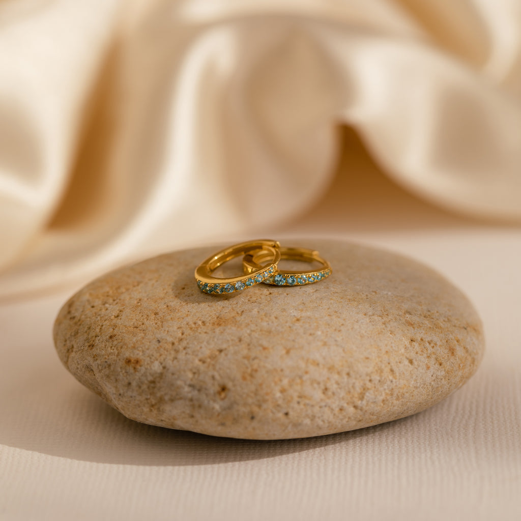 The Vera Pave Aquamarine Huggies, two gold hoops with sparkling blue gemstones, rest on a smooth stone against a soft cream fabric—an elegant match for your favorite gemstone earrings.