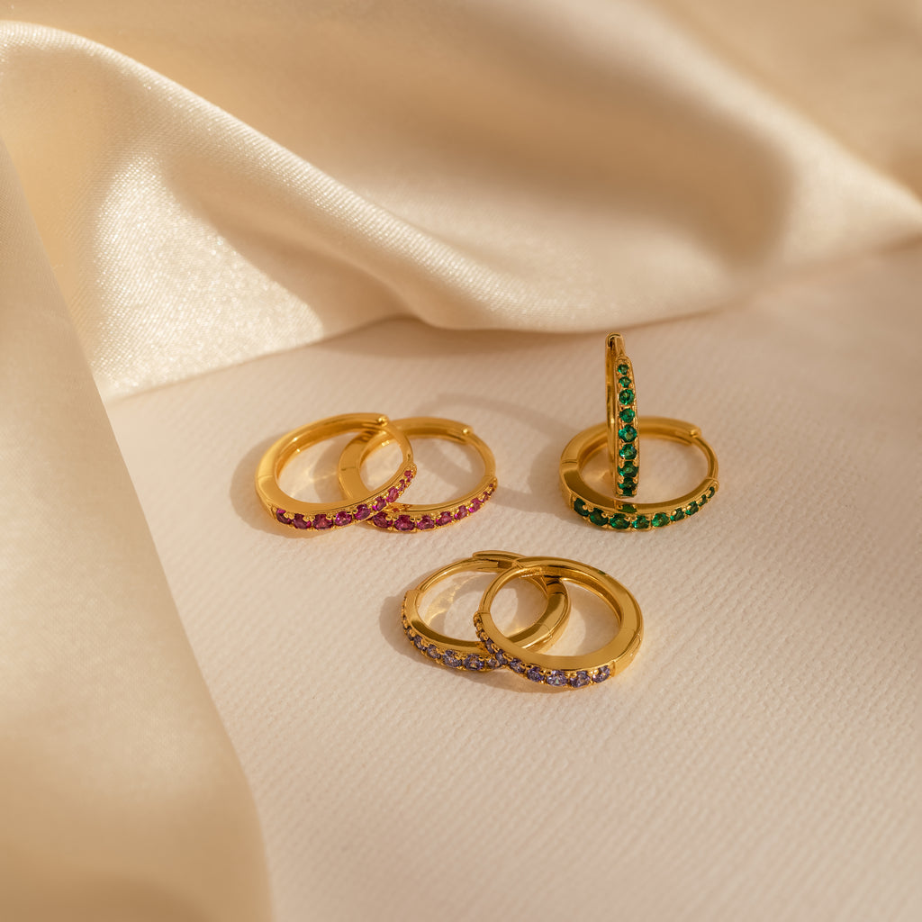 Vera Pave Birthstone Huggies, featuring vibrant gemstones, are showcased on a soft beige satin fabric for a colorful and elegant look.