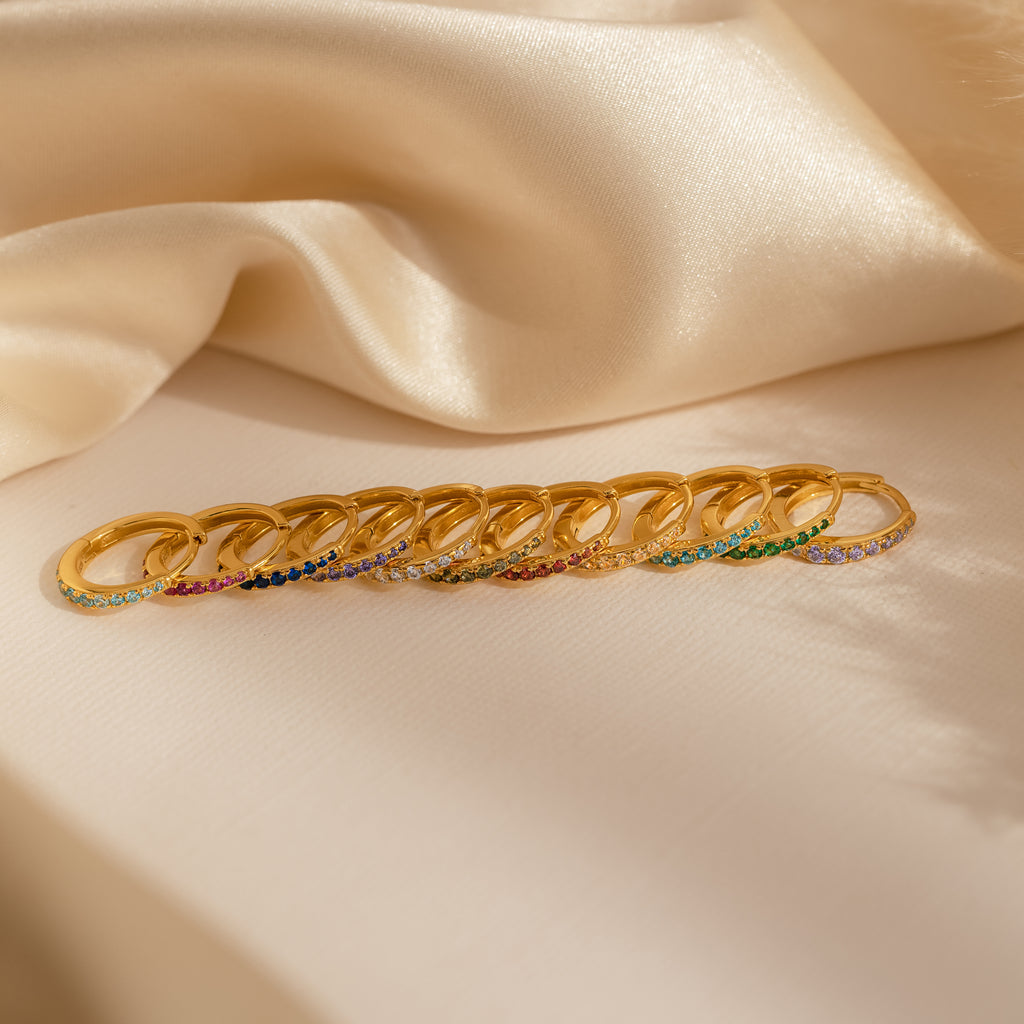 Ten vibrant earrings, including Vera Pave Birthstone Huggies, are displayed on cream-colored satin fabric.