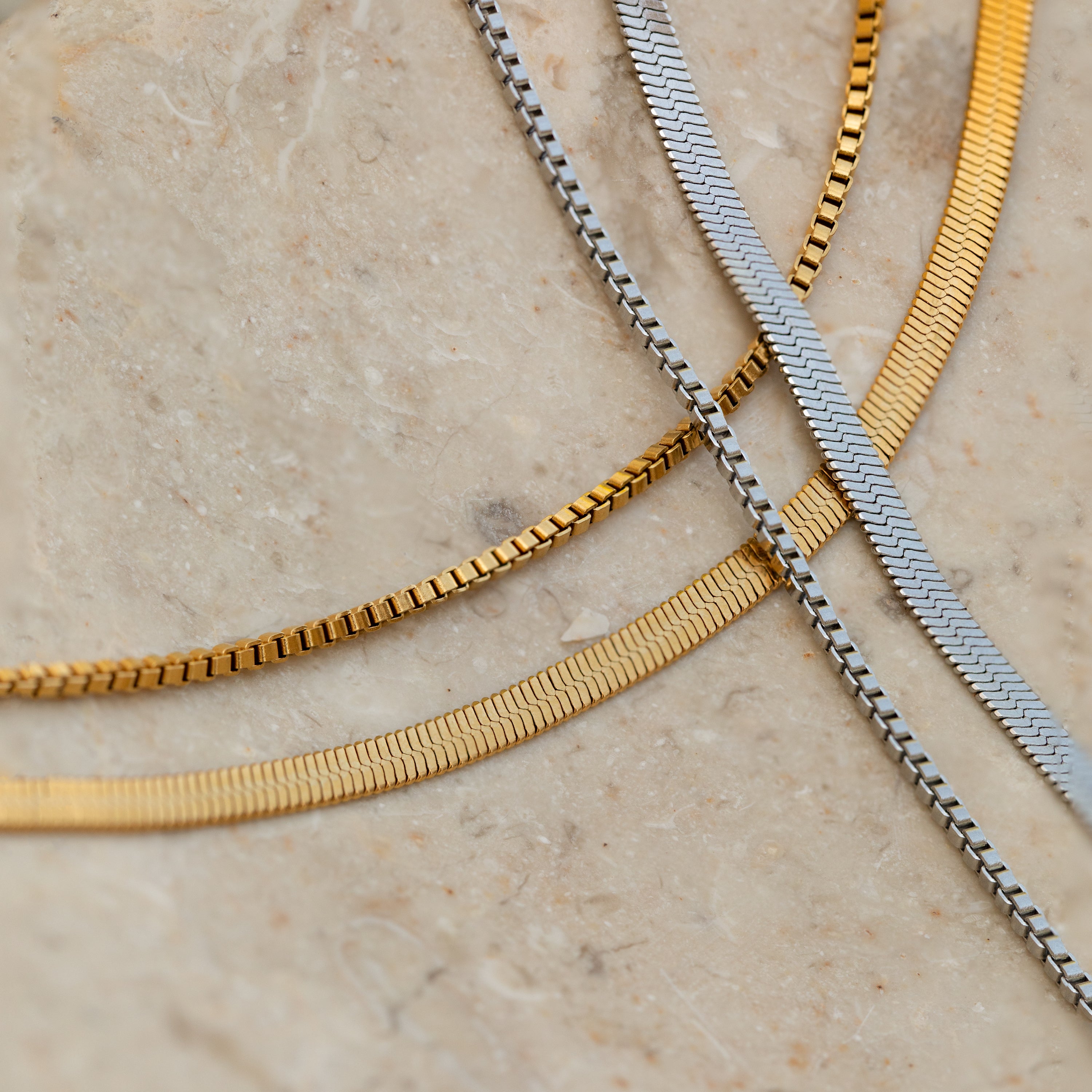 The Herringbone Duo Chain Necklace in Silver is displayed on a beige stone surface, showcasing its sleek, classic style for a minimalist necklace look.