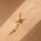 A pair of delicate gold ear climbers on a textured beige surface, lit by dramatic diagonal sunlight that casts soft shadows.