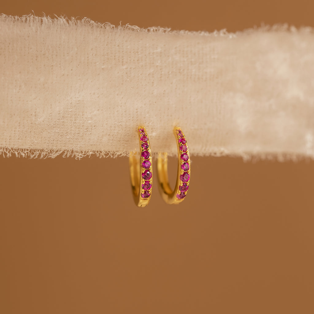 Vera Pave Birthstone Huggies, gold hoop earrings adorned with small pink gemstones, are displayed on textured cream fabric with a brown background.