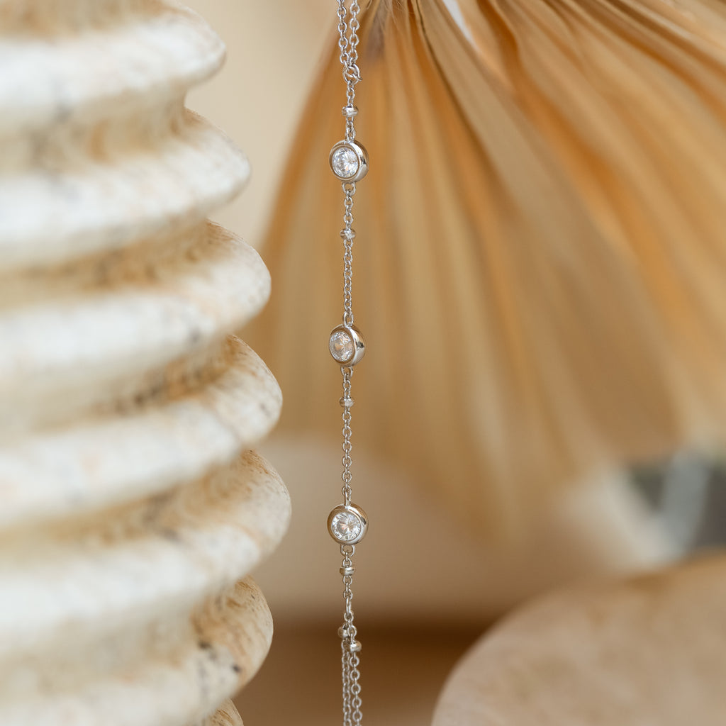 The Diamond Bezel Hand Chain in Sterling Silver features small round diamonds, showcased on a neutral, textured background.
