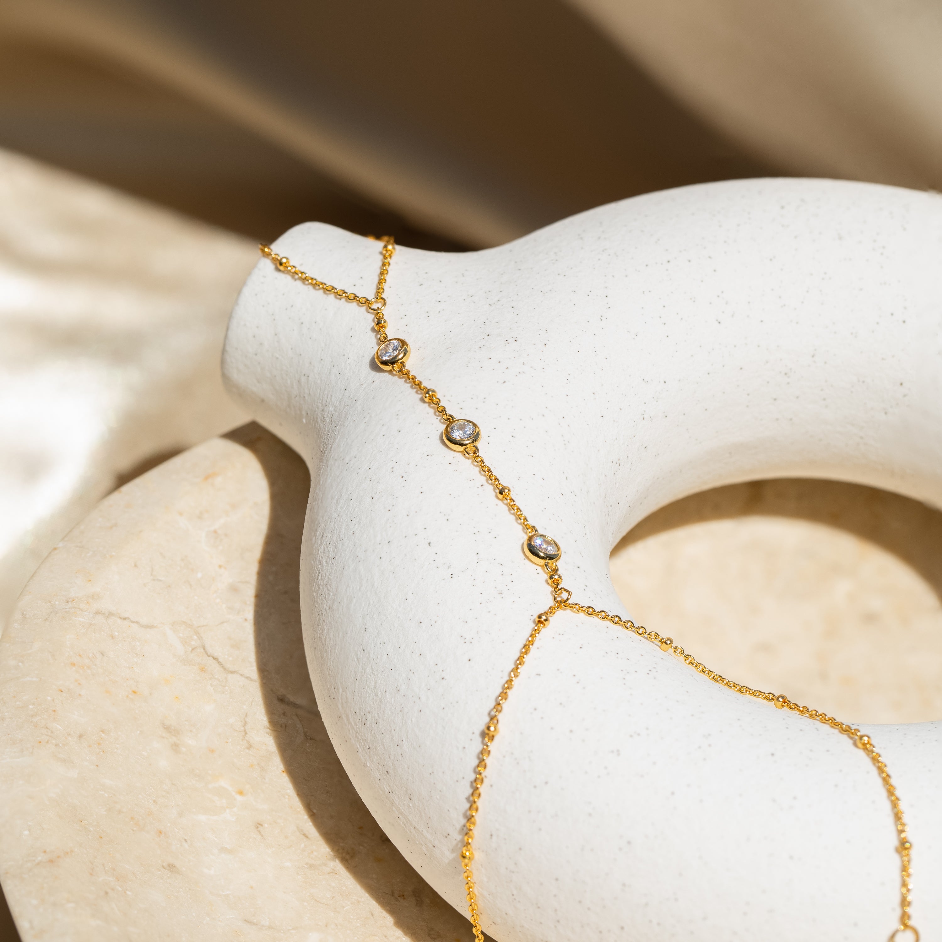 The Diamond Bezel Hand Chain, a gold chain bracelet with three small gemstones, is draped on a white round ceramic vase.