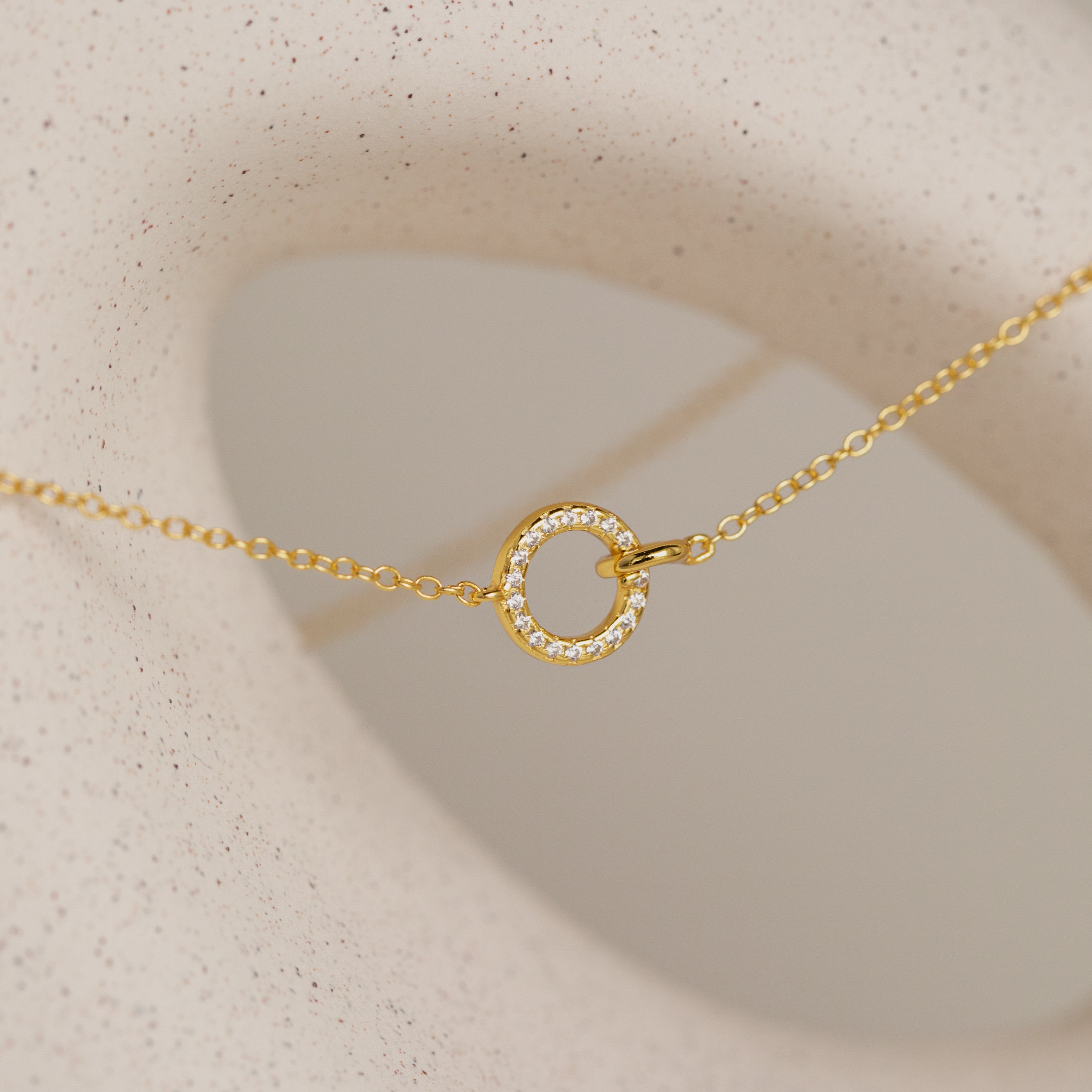 Pave Interlocking Necklace: A gold bridal necklace with a small, diamond-studded circle pendant displayed on a textured cream surface—an elegant unity jewelry piece.