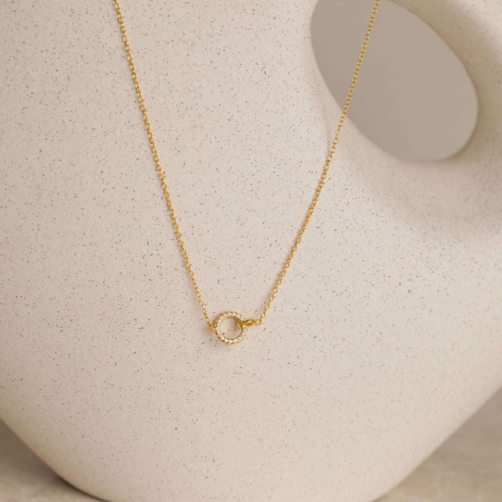 The Pave Interlocking Necklace in 18K Gold features a small circle pendant, elegantly displayed against a cream ceramic vase—ideal as bridal jewelry or a meaningful unity necklace.