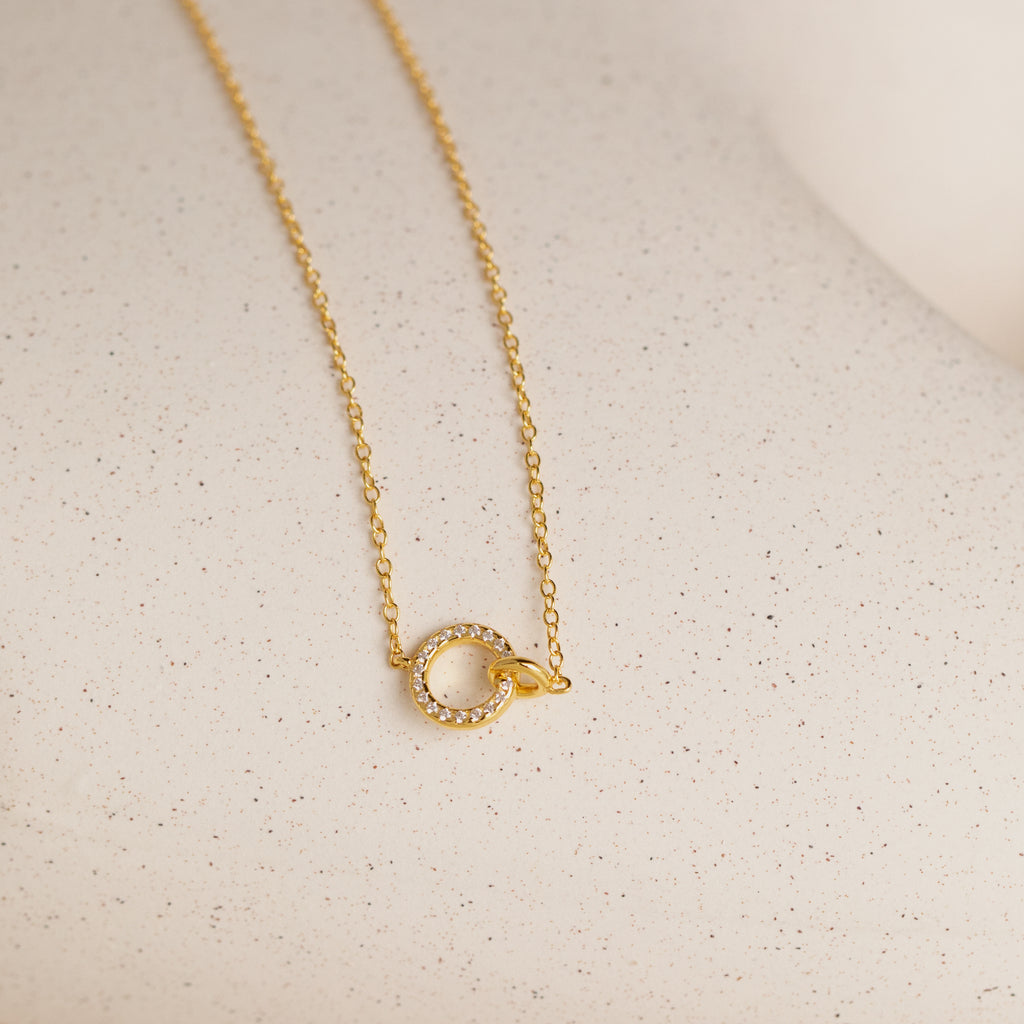 The Pave Interlocking Necklace in 18K Gold features a delicate chain with a small, circular gemstone pendant—perfect as elegant bridal jewelry on a light surface.