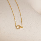 The Pave Interlocking Necklace in 18K Gold features a delicate chain with a small, circular gemstone pendant—perfect as elegant bridal jewelry on a light surface.