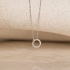 The Pave Interlocking Necklace in Silver features a sparkling circular pendant on a neutral textured surface—an elegant keepsake and perfect choice for bridal jewelry.
