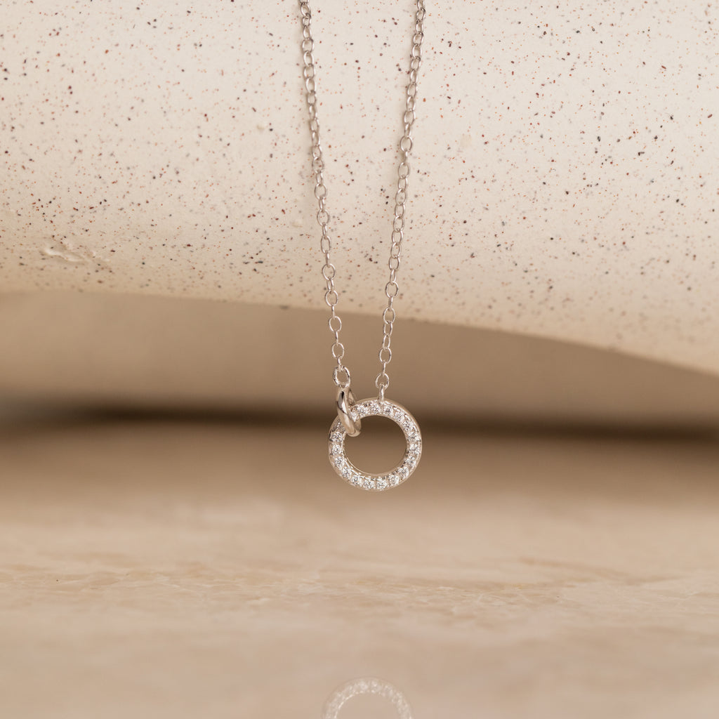 The Pave Interlocking Necklace is an elegant silver necklace featuring a small circular diamond-studded pendant, showcased against a neutral textured background.