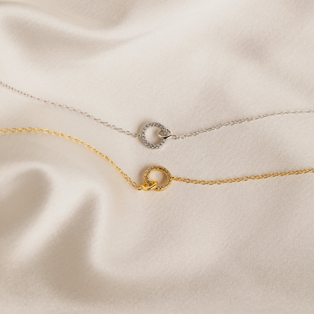 A delicate silver chain necklace with a circular pendant rests on silky beige fabric—an elegant Pave Interlocking Necklace in Silver, perfect as bridal jewelry or a timeless keepsake.