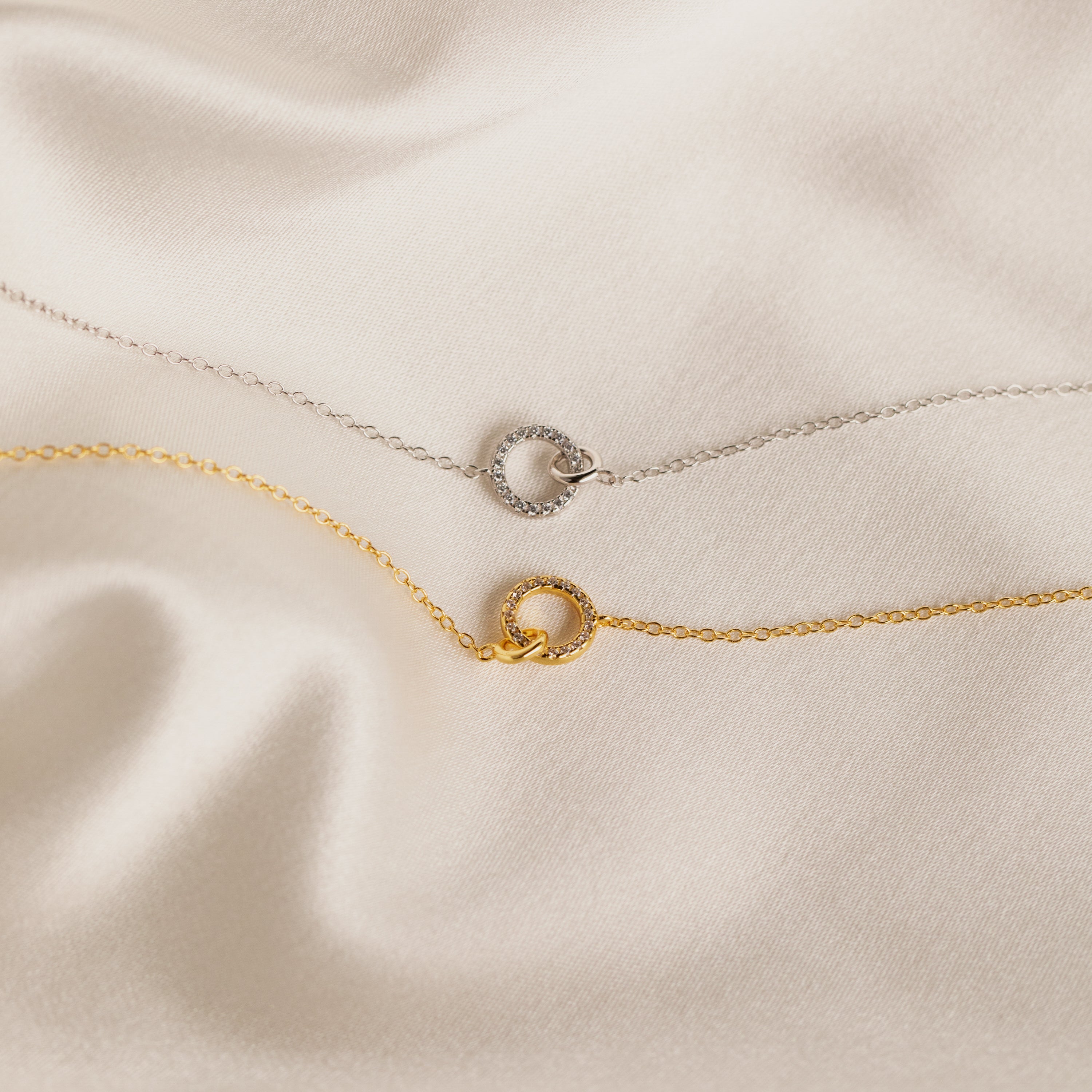 A delicate silver chain necklace with a circular pendant rests on silky beige fabric—an elegant Pave Interlocking Necklace in Silver, perfect as bridal jewelry or a timeless keepsake.