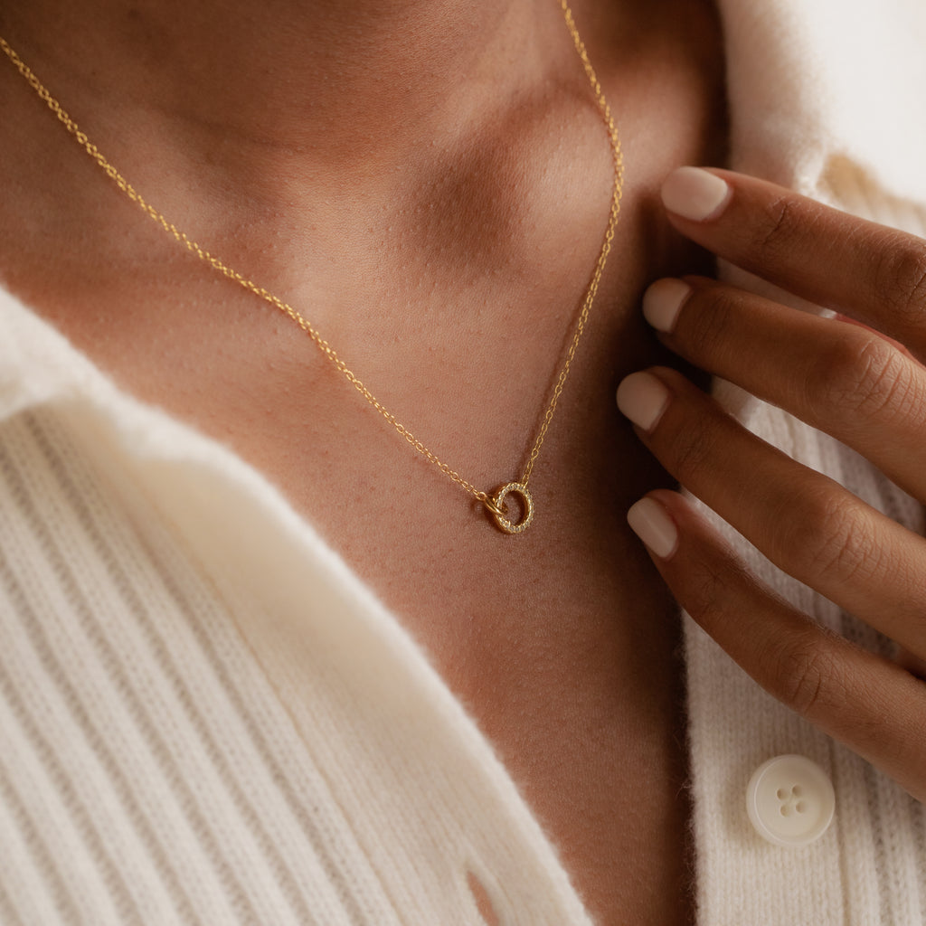 Close-up of the Pave Interlocking Necklace—a unique unity piece featuring a circular pendant—worn over a cream ribbed shirt with a button.