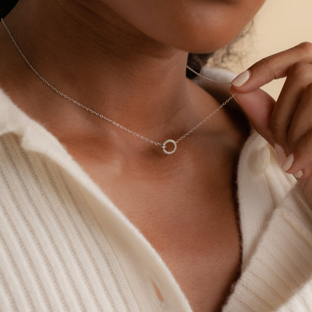 A woman wears the Pave Interlocking Necklace—a delicate silver jewelry piece with a small circular pendant—holding it gently near her collar.